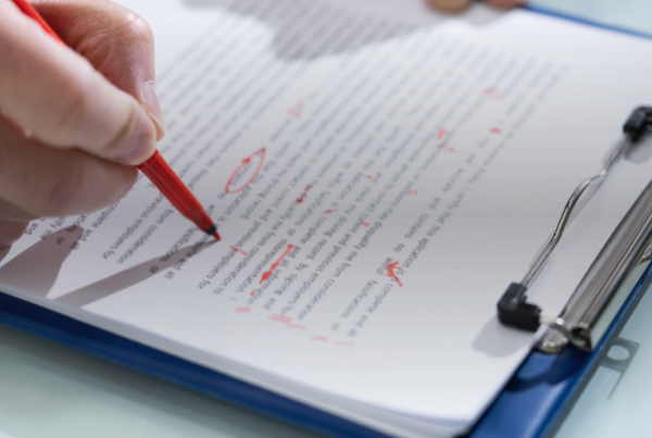 Mão com caneta vermelha corrigindo um texto em uma prancheta