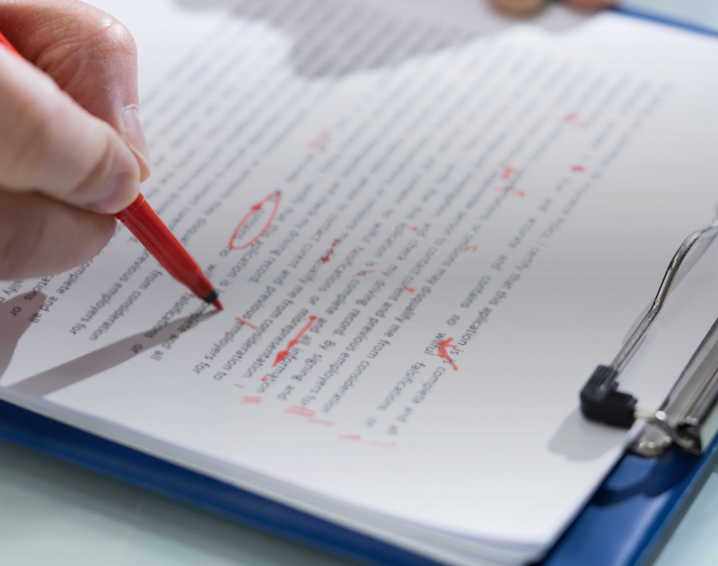 Mão com caneta vermelha corrigindo um texto em uma prancheta